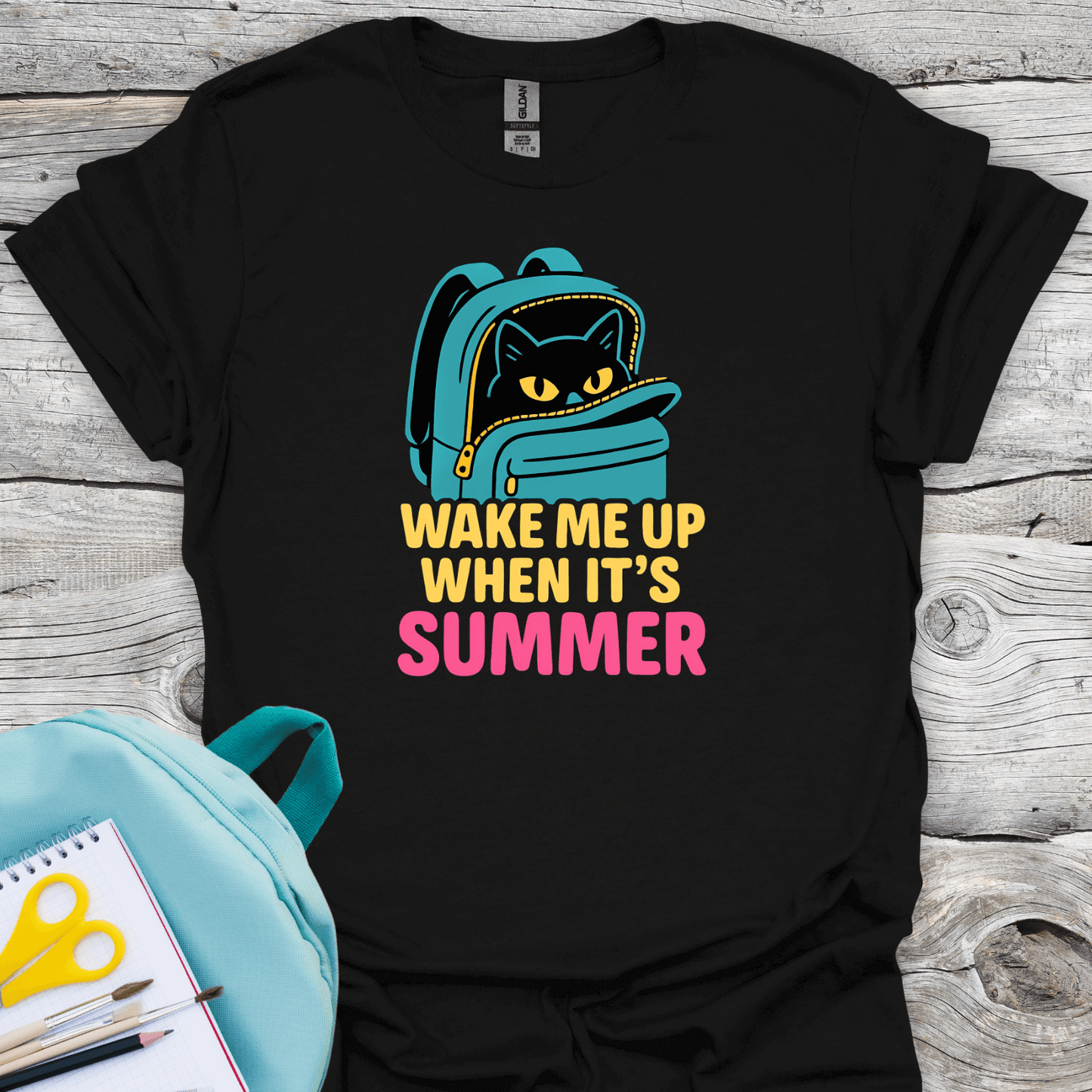 Black t-shirt with a cat in a backpack graphic and 'Wake Me Up When It's Summer' text on a wooden surface.