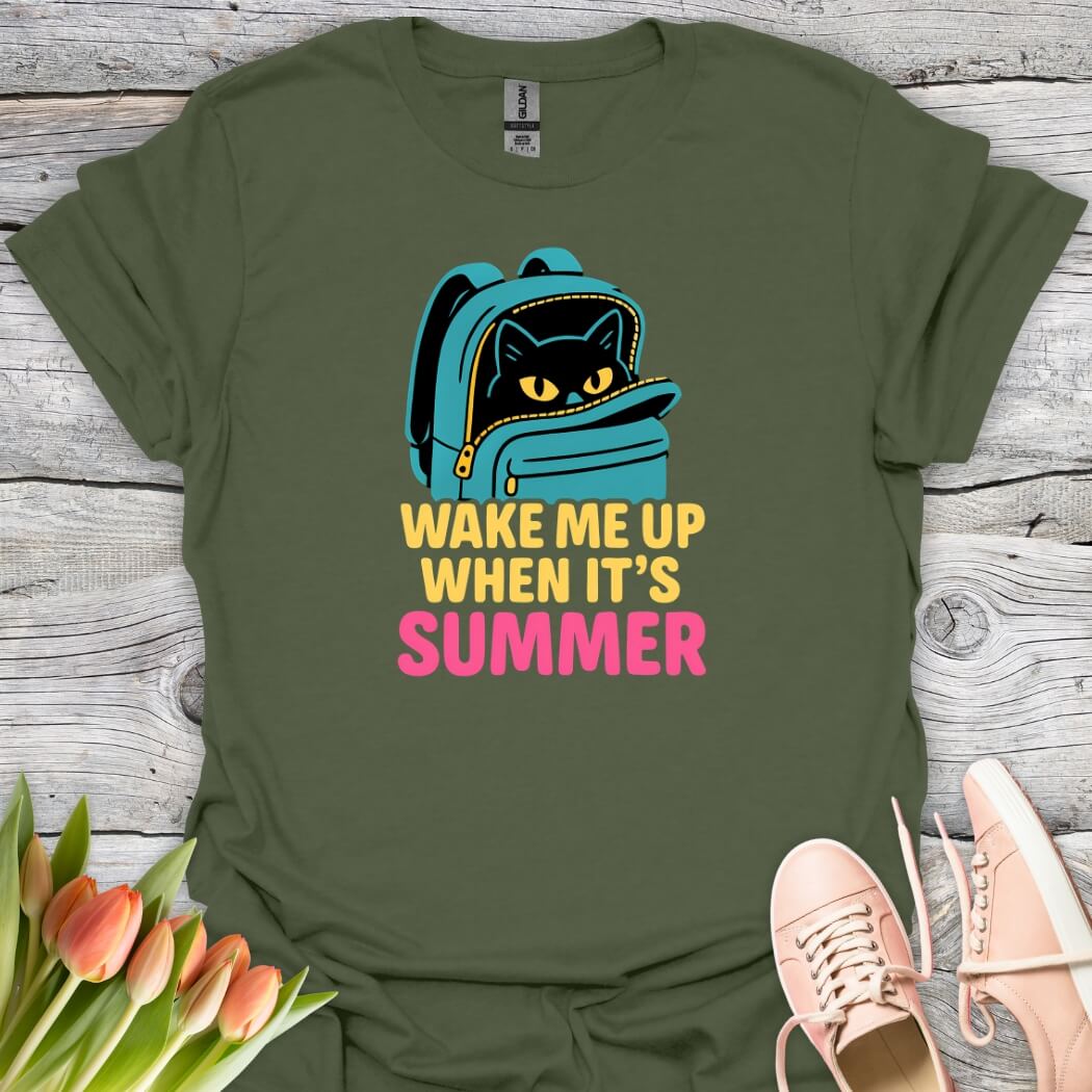 Green t-shirt with a cat in a backpack graphic and 'Wake Me Up When It's Summer' text on a wooden surface with flowers and shoes.